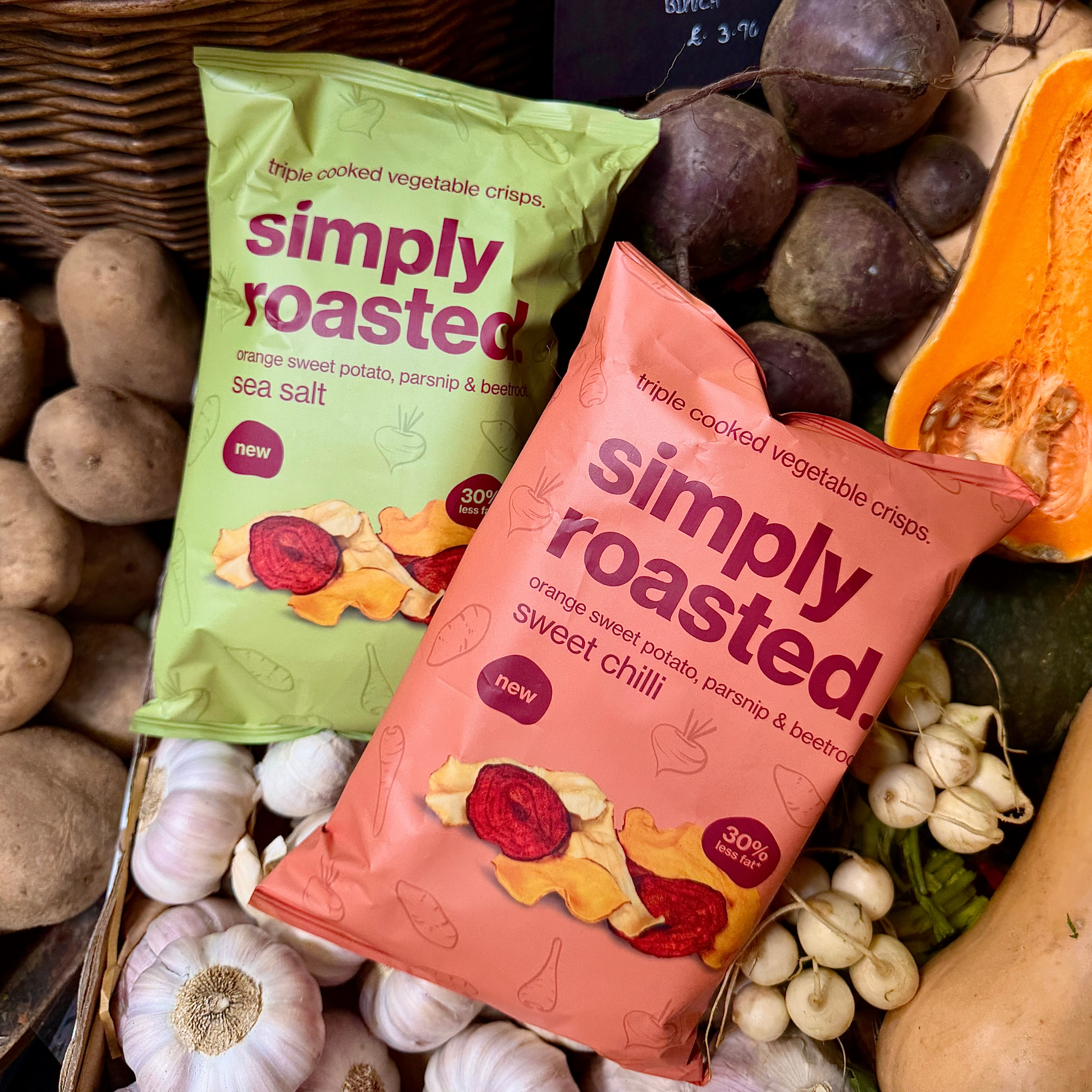 Two bags of Simply Roasted vegetable crisps displayed among their raw ingredients, including parsnips, beets, garlic, potatoes, and a squash. The bag in the foreground is orange-pink and labeled 'simply roasted' 'sweet chilli', while the bag in the background is light green and labeled 'simply roasted' 'sea salt'.
